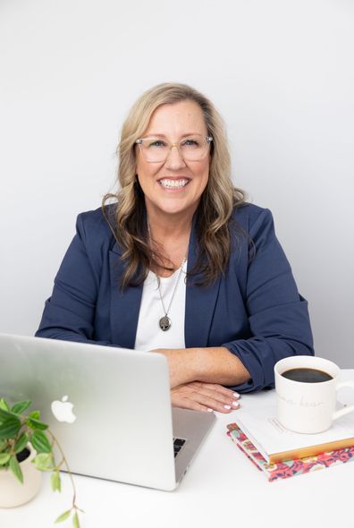 Woman smiling at a desk with a laptop, coffee mug, and notebook in a bright office.