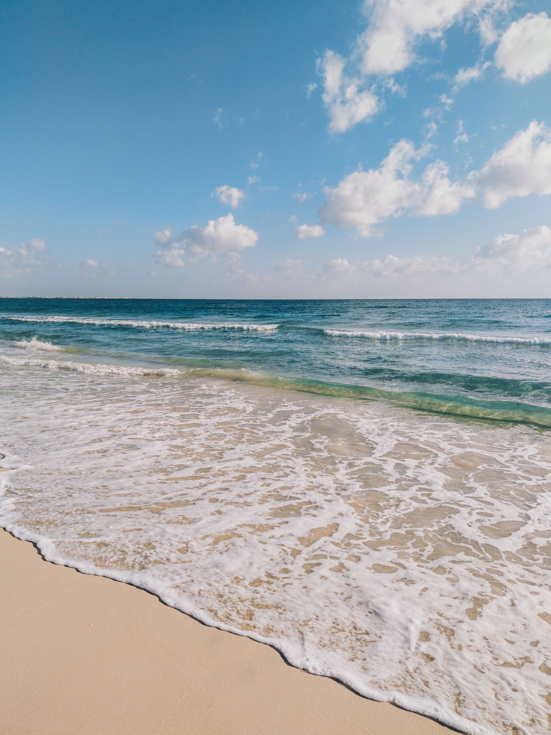 Sandy beach with foamy waves, turquoise sea, and a blue sky with scattered clouds