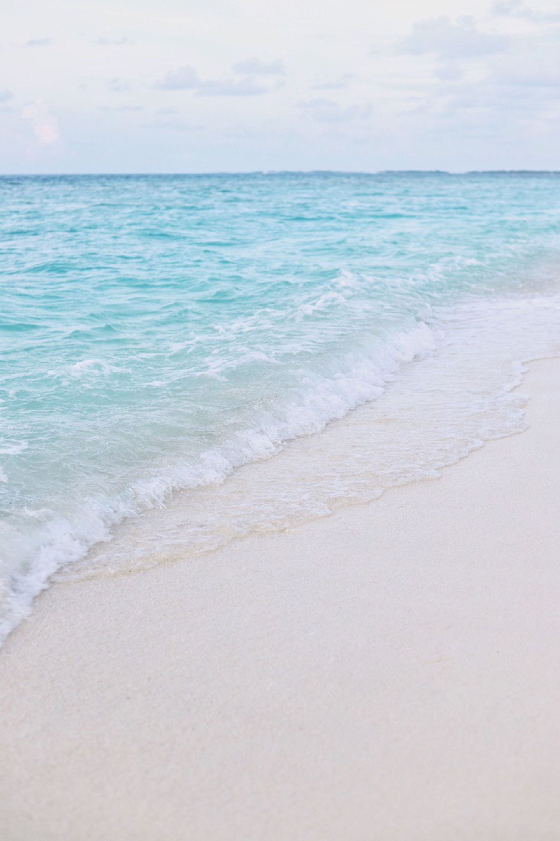 Turquoise ocean waves washing onto a white sandy beach under a pale sky