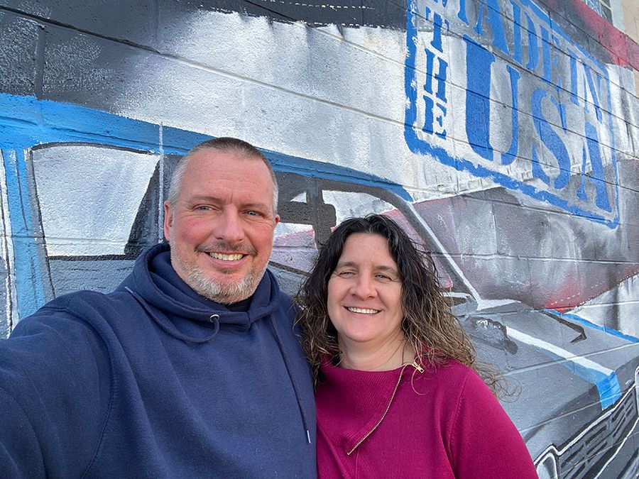 Two smiling people in hoodies posing in front of a mural with large blue “USA” letters. | Tinley Auto Repair & Towing Inc