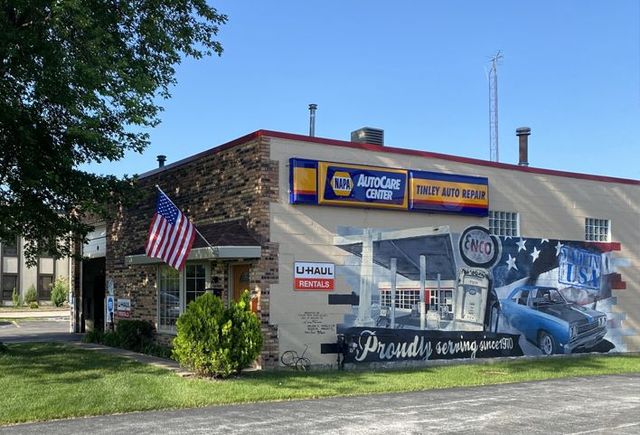 Auto service shop exterior with American flag, blue car mural, and signs under a clear sky | Tinley Auto Repair & Towing Inc