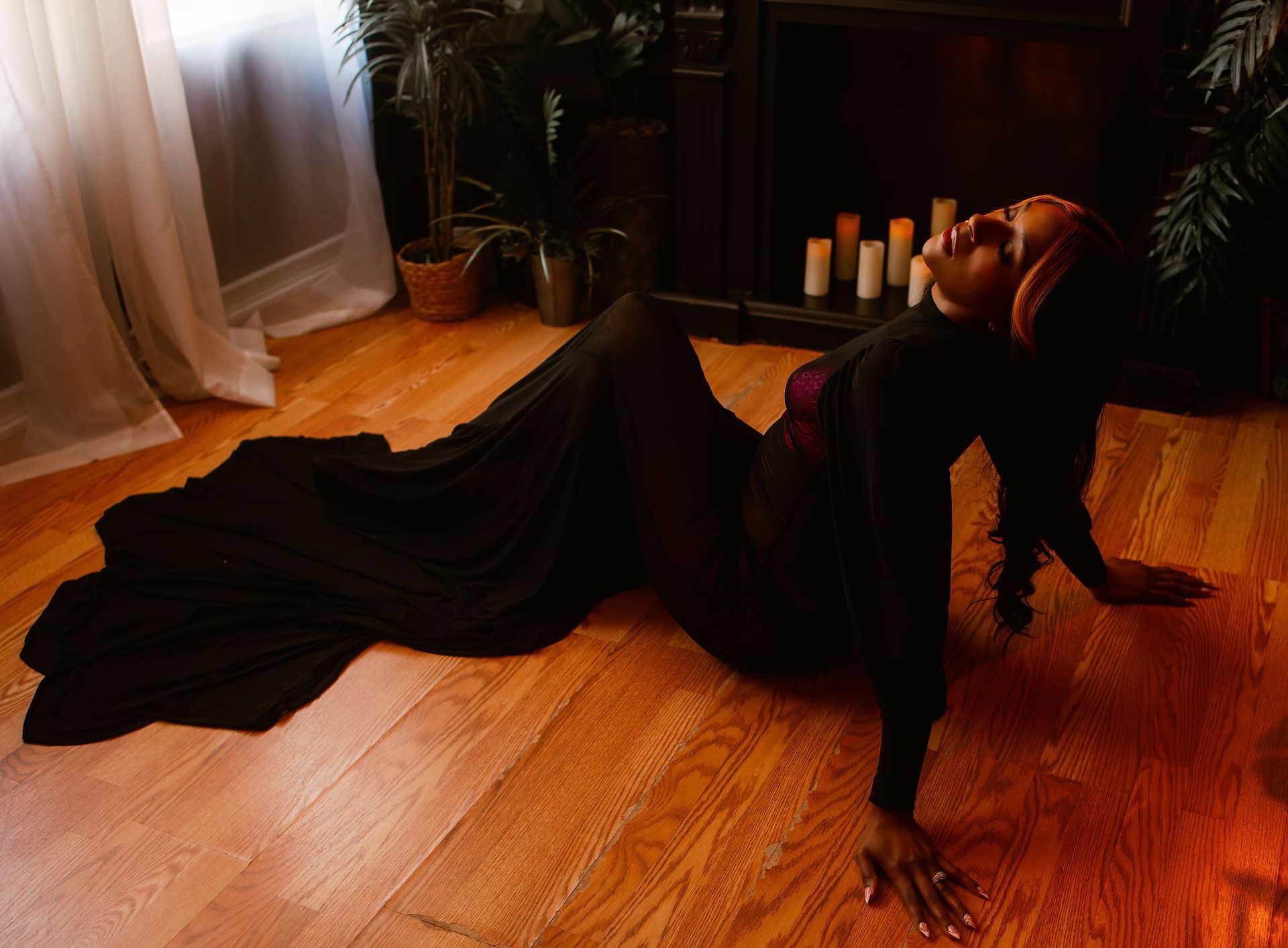 Person in a black dress posing on a wooden floor in a dim room with candles and plants