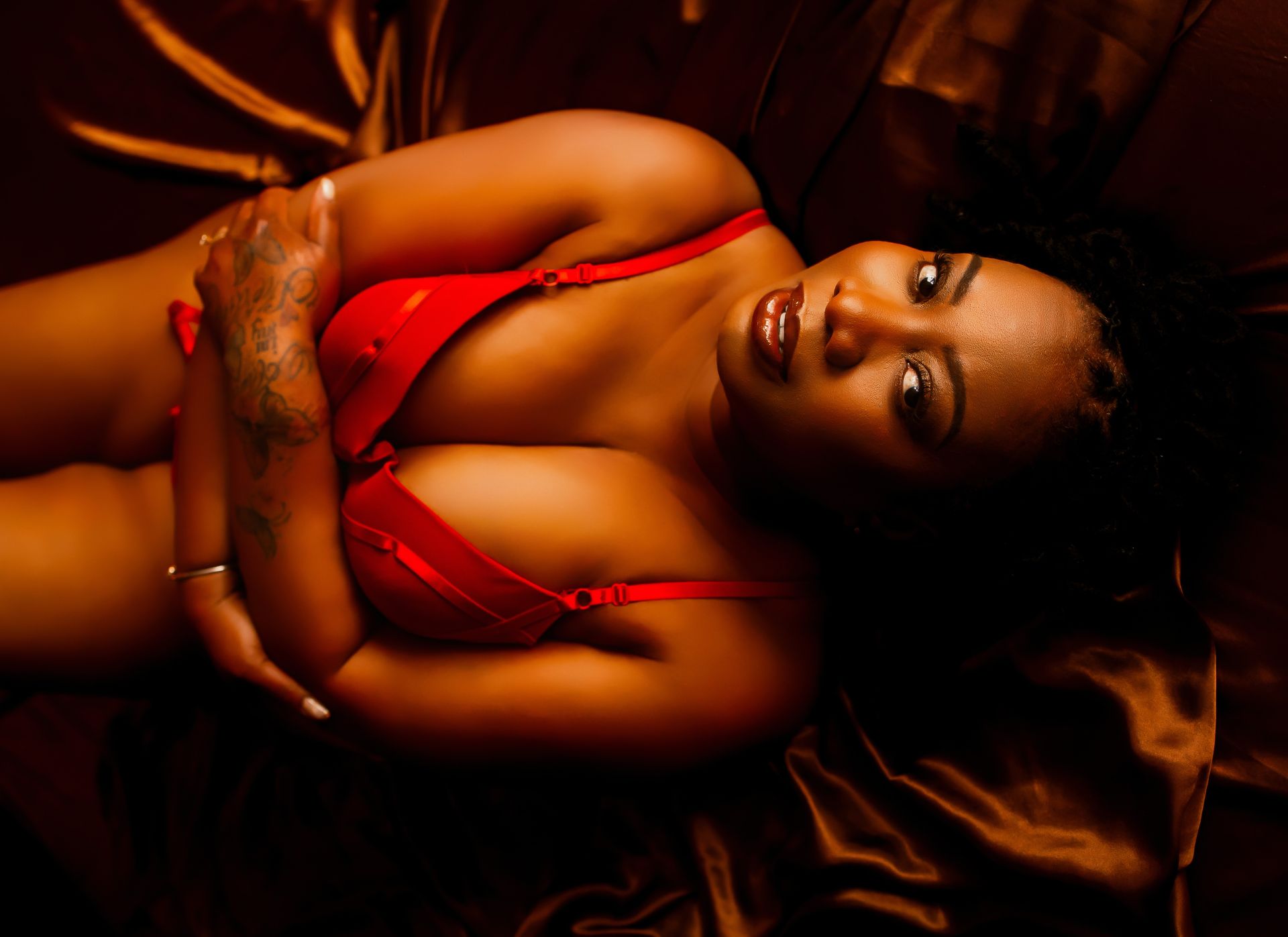 Person in a red outfit reclining on dark satin, looking at the camera.