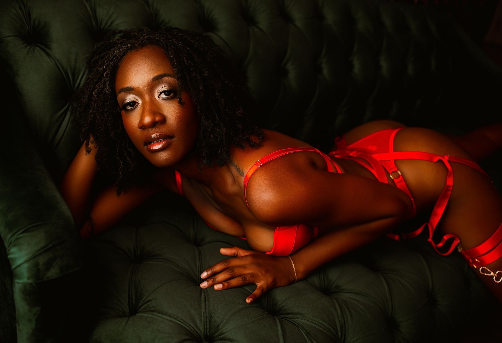 Person posing on a dark green tufted sofa, wearing red lingerie in a low-lit setting