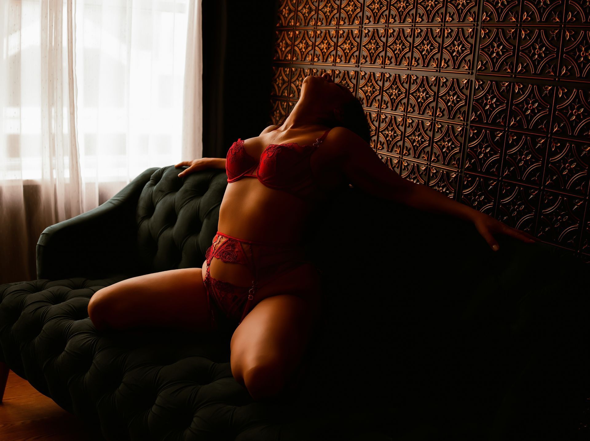Woman reclining on a dark sofa in red lingerie by a window, with patterned wallpaper behind her.