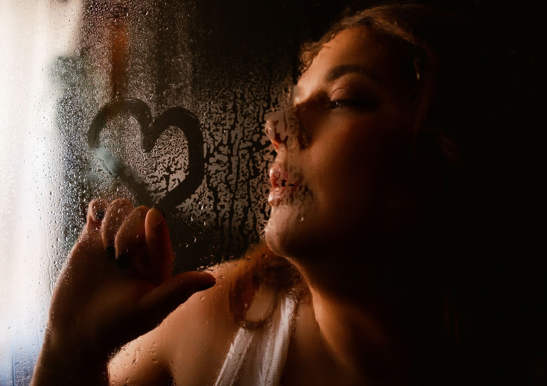 Woman seen through fogged glass, touching a heart drawn on the window in warm light.