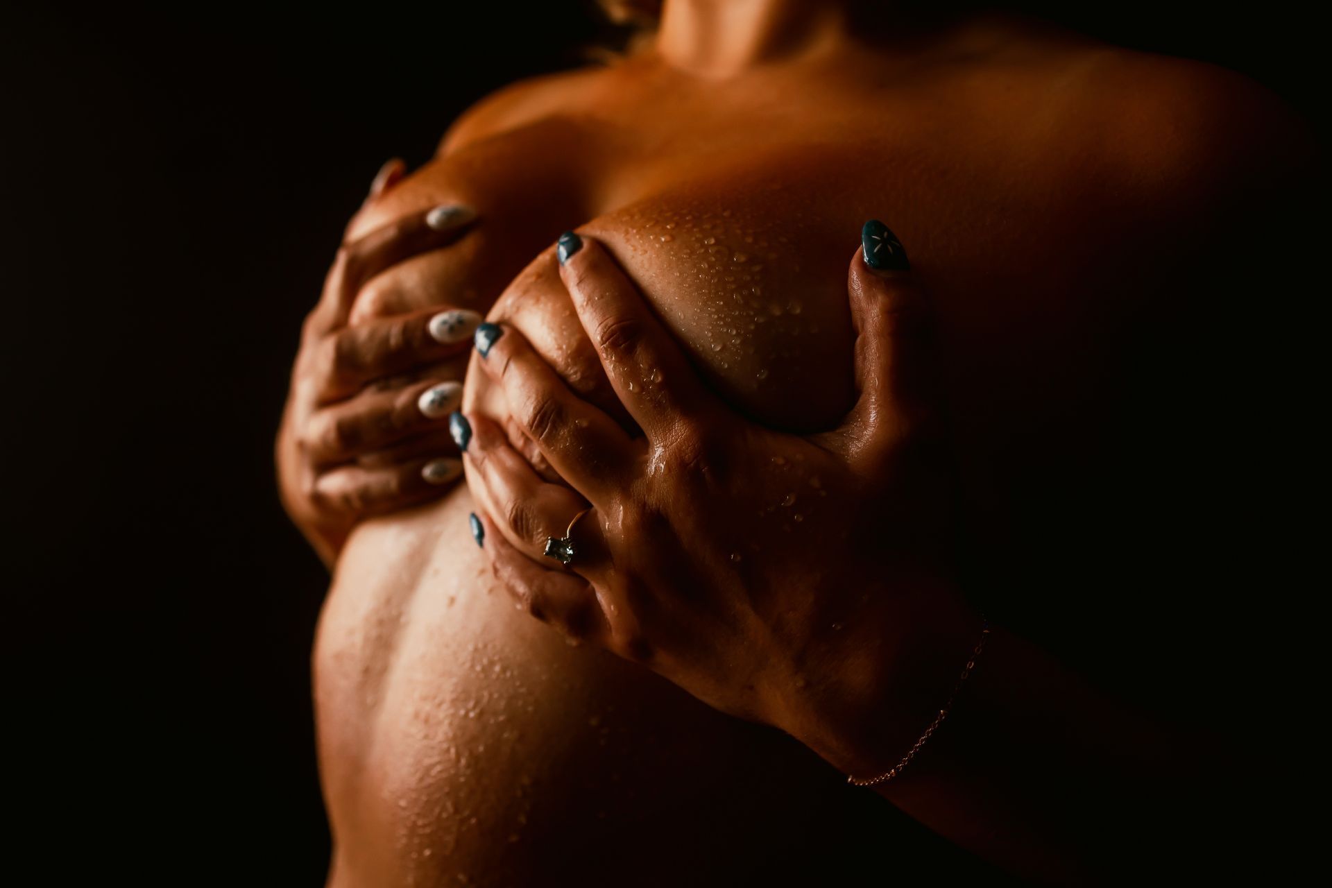 Hands with dark polished nails caress a bare, wet back in warm low light.