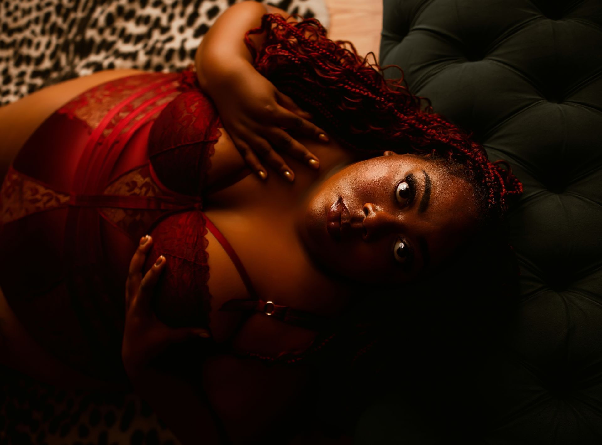 Woman reclining on a dark tufted couch in red lace lingerie, with curly red hair and dramatic makeup.