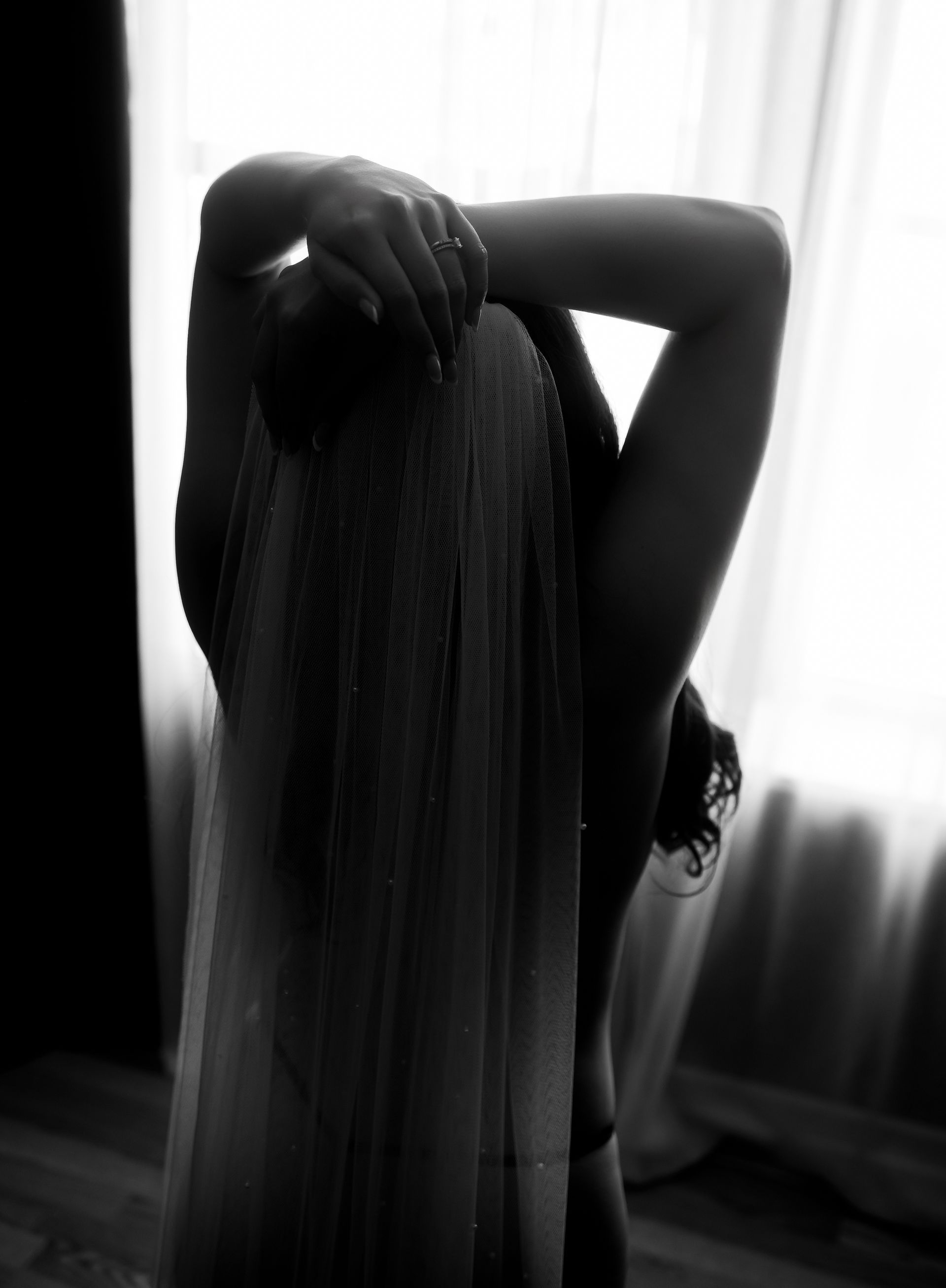 Silhouetted person with long hair standing by a bright window, hands raised to their head