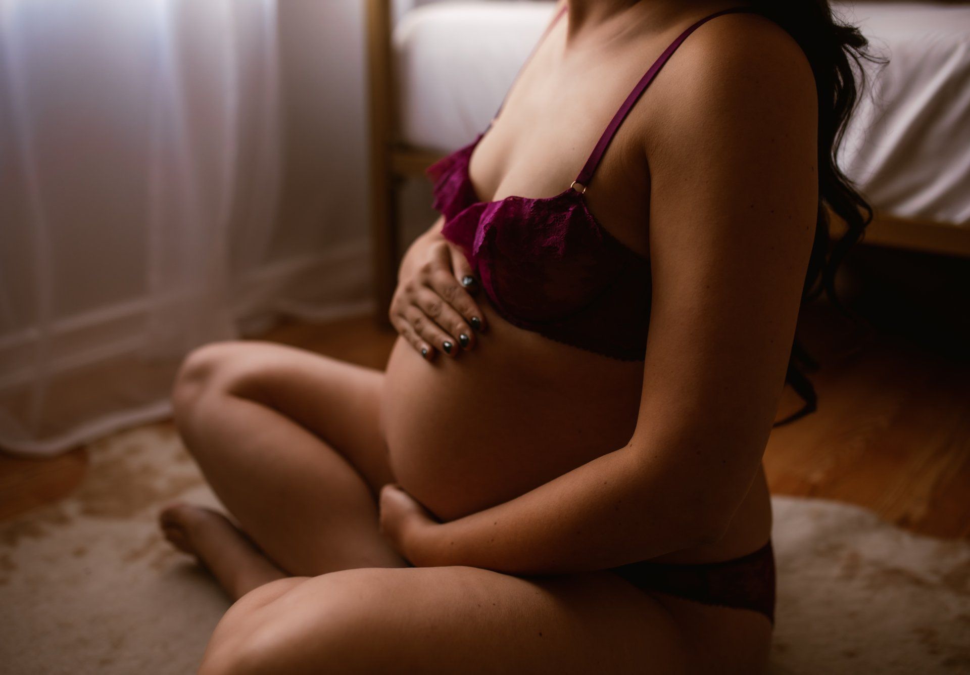 A pregnant woman in lingerie is sitting on the floor holding her belly.