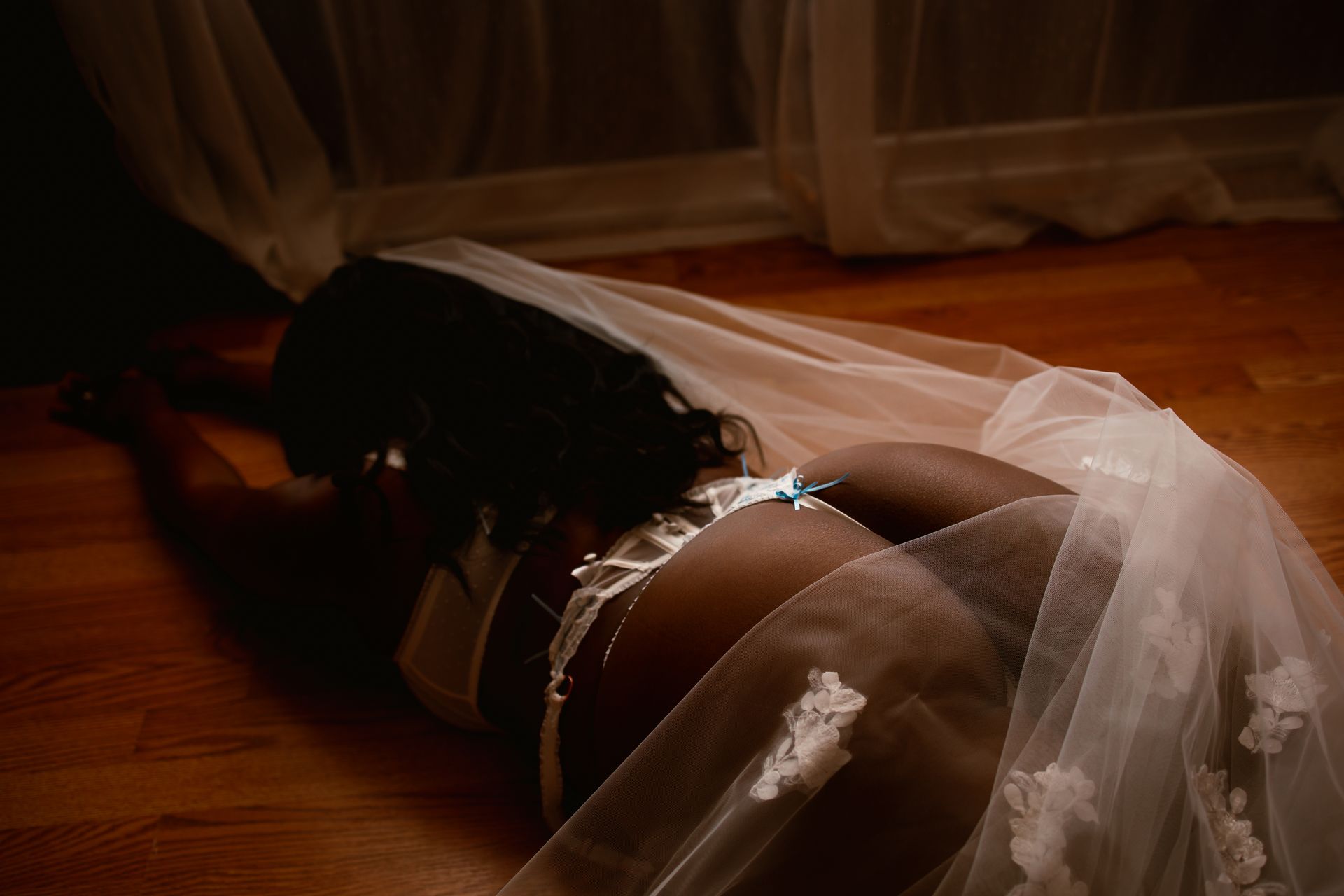 A person lying on a wooden floor beneath a sheer white veil in warm, dim light
