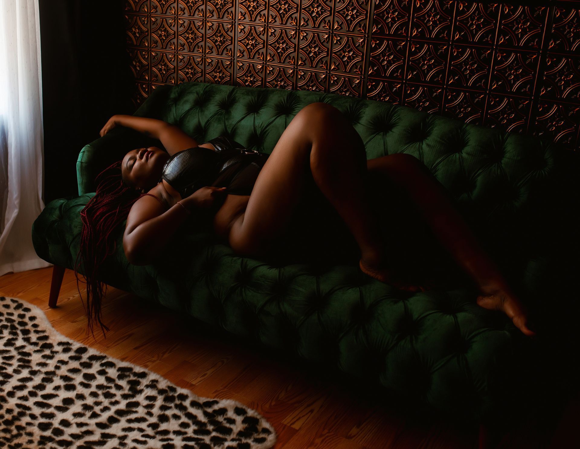 Person reclining on a dark green velvet sofa in a dimly lit room with a patterned rug and sheer curtains.
