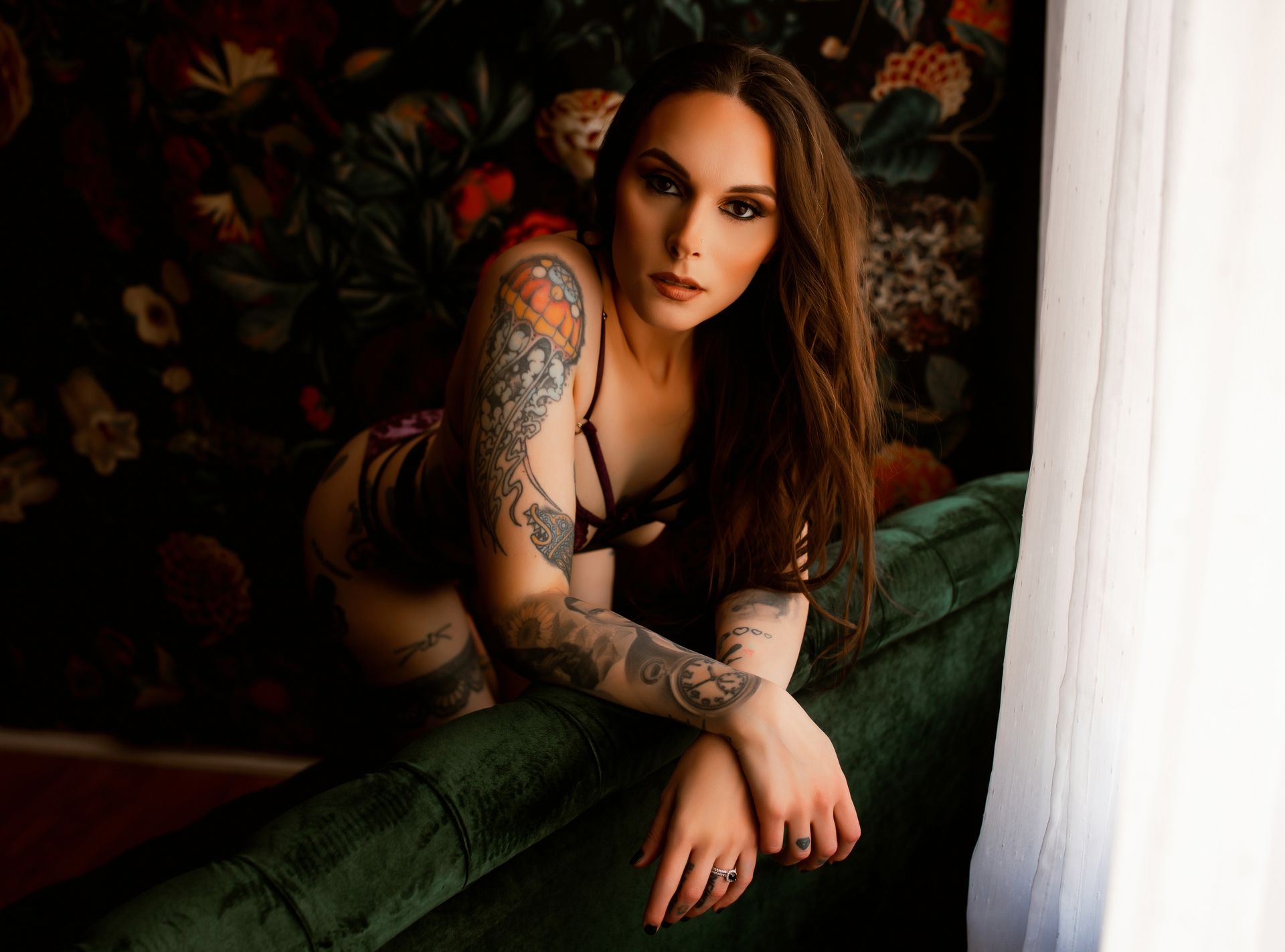 A woman with tattoos is sitting on a green couch in front of a window.