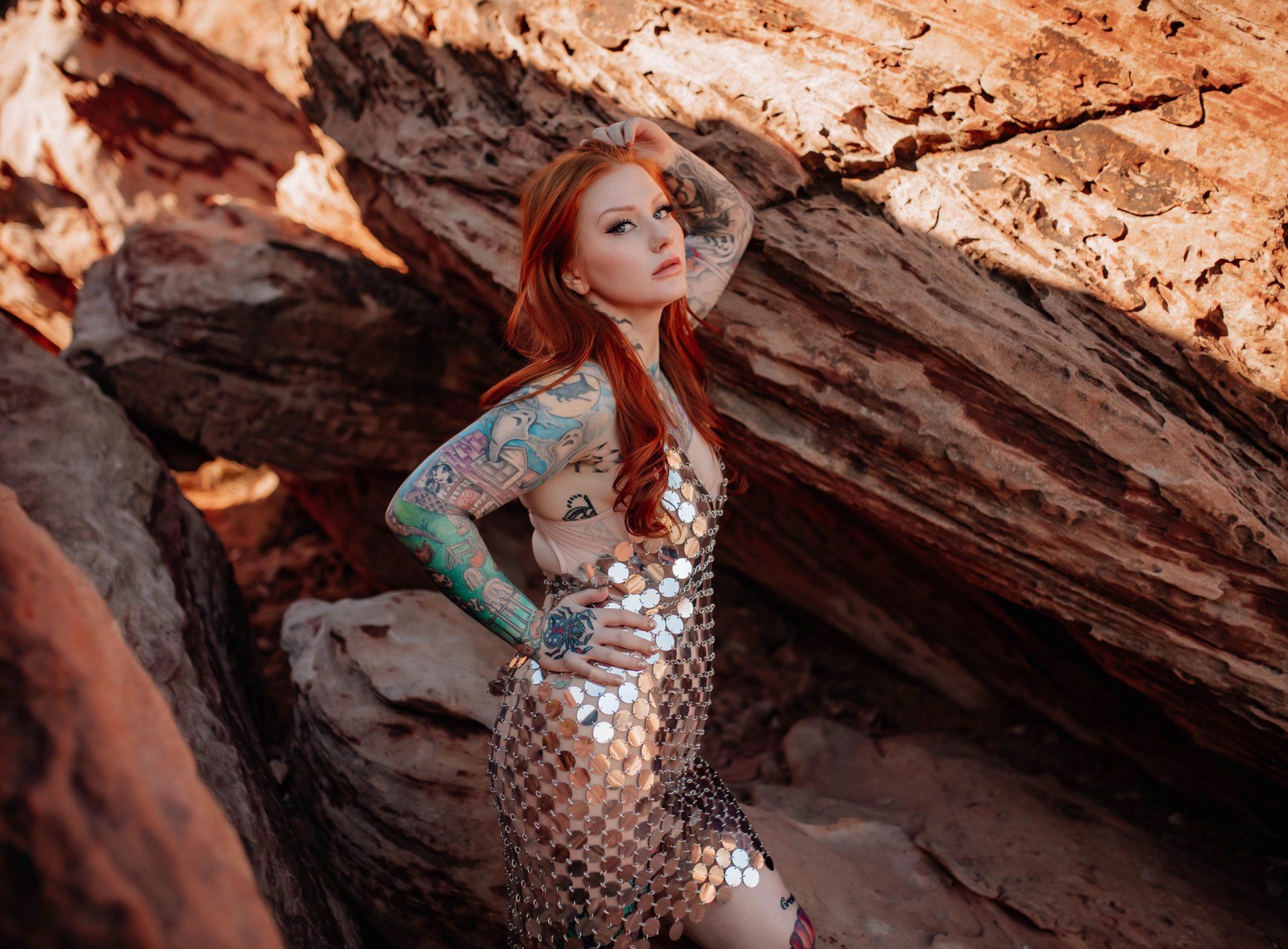 A woman with red hair and tattoos is standing in front of a rock formation.
