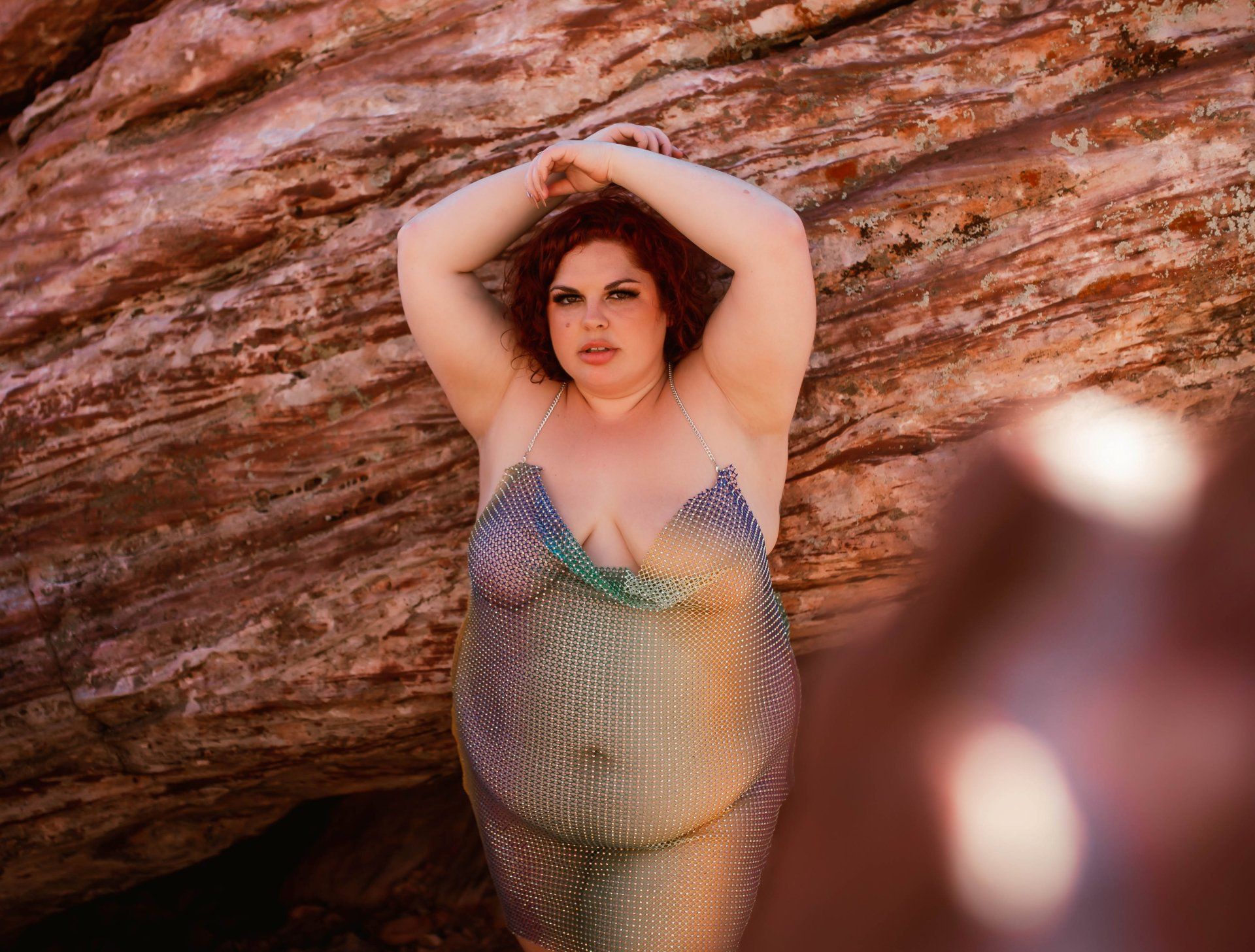 A woman in a sheer dress is standing in front of a large rock.