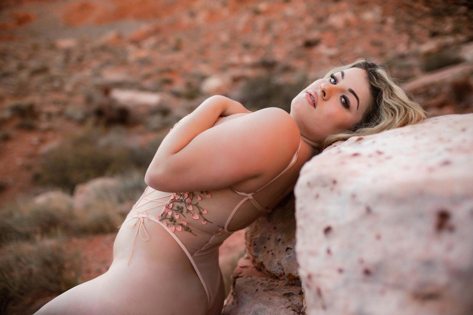 Woman reclining outdoors on rocks in a beige lace bodysuit, looking toward the camera.