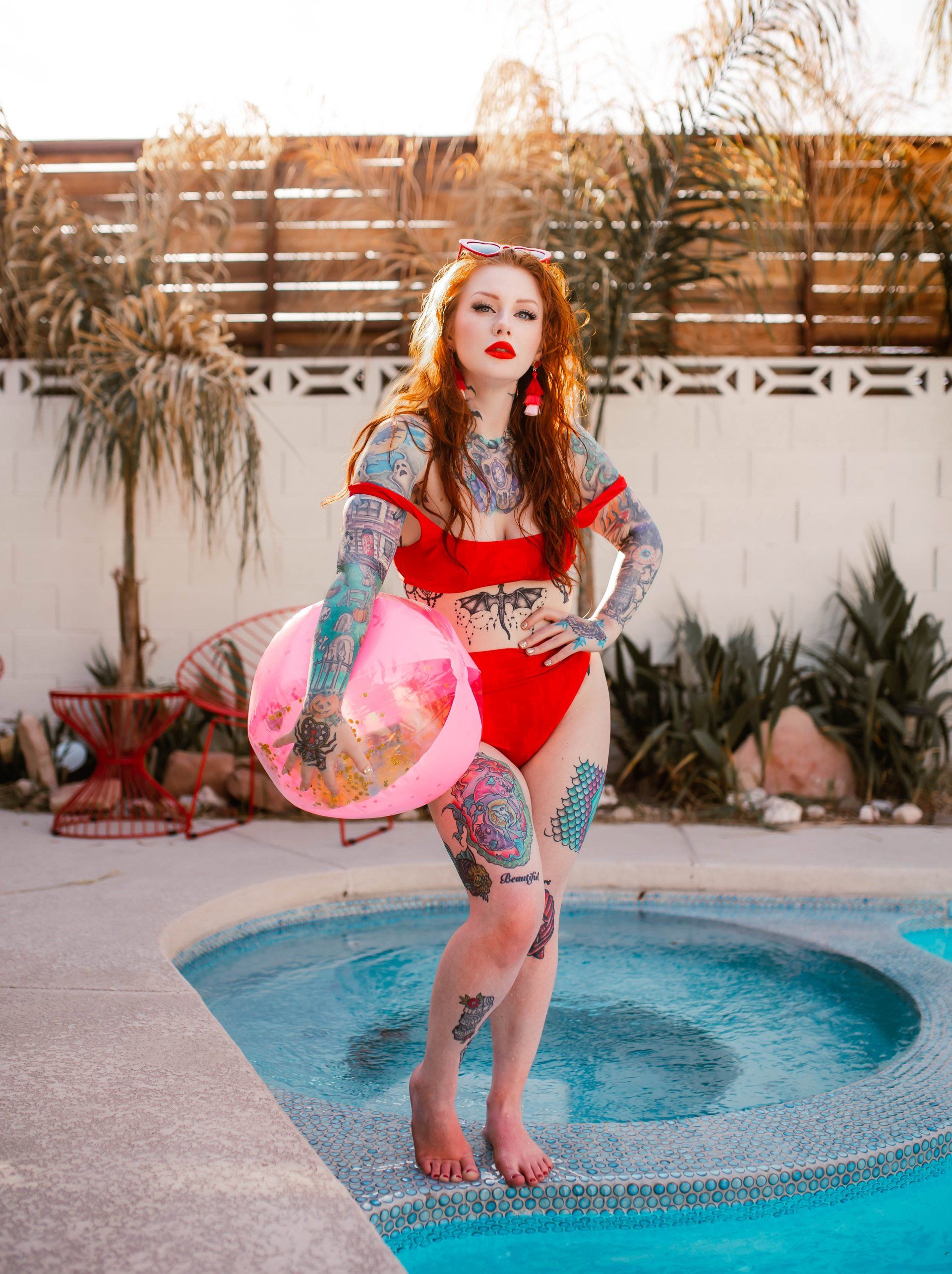 A woman in a red bikini is standing next to a swimming pool holding an inflatable ball.