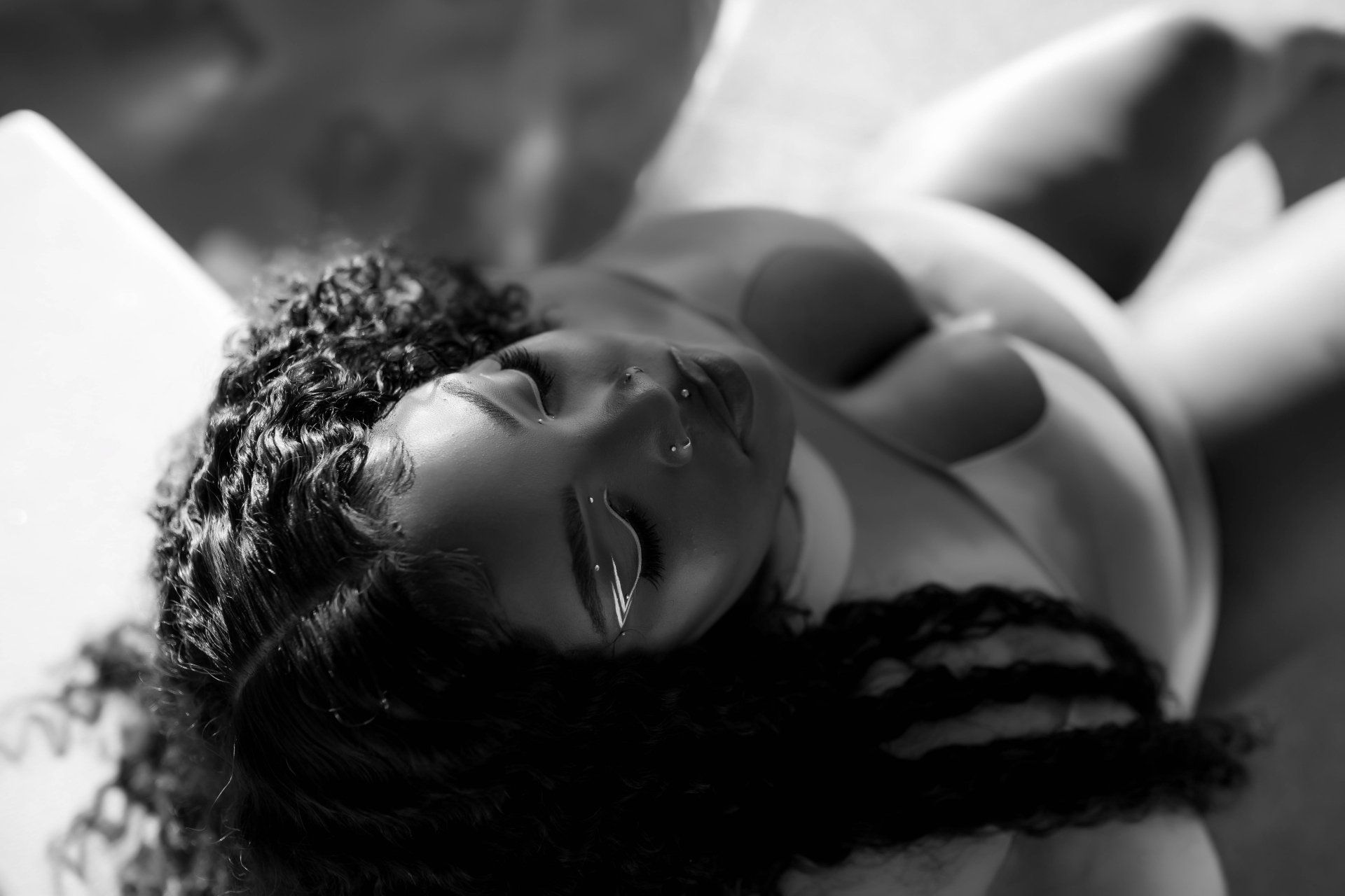 Black-and-white portrait of a person reclining with curly hair, wearing a light top, looking toward the camera