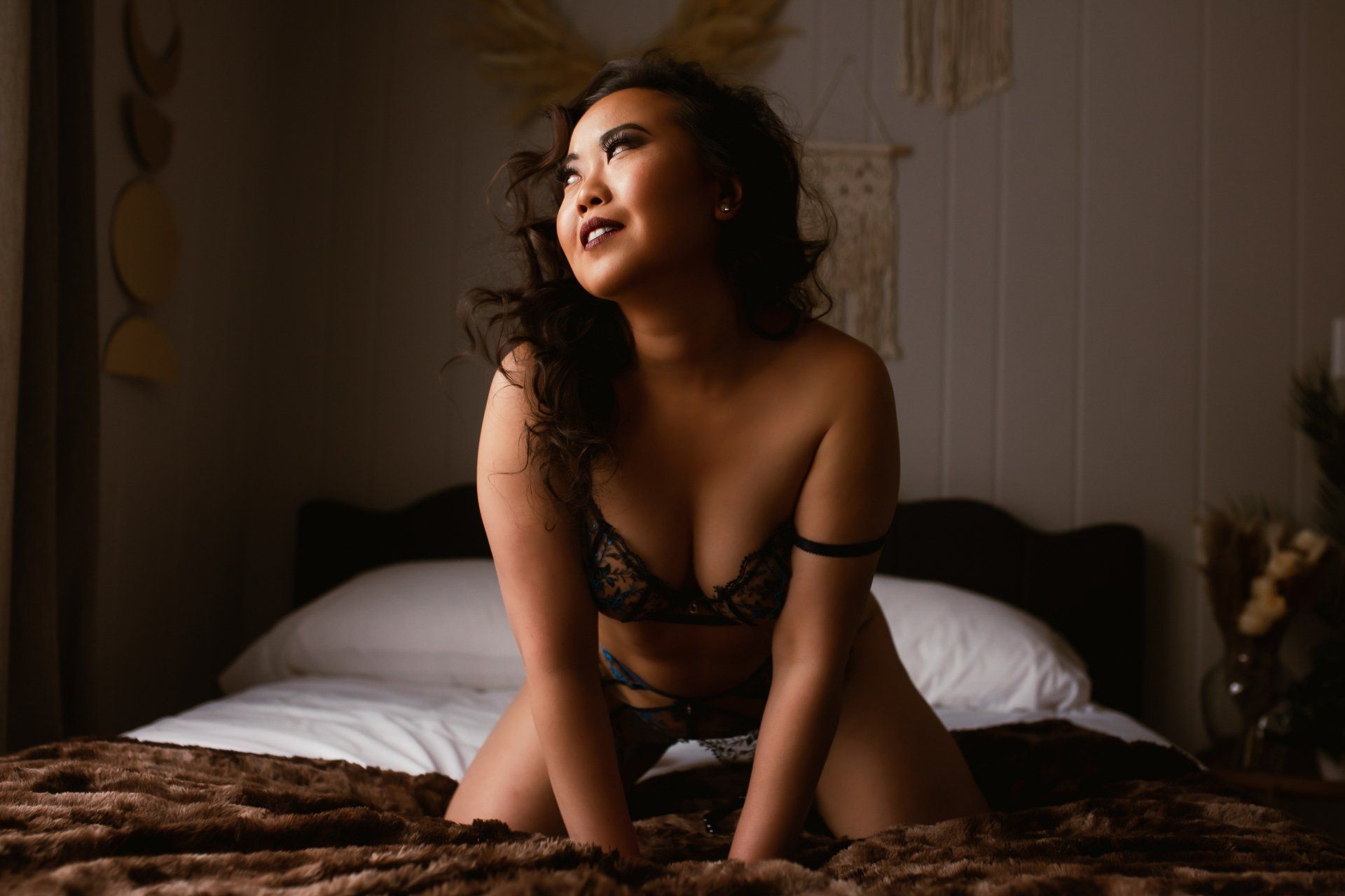 Woman in lingerie sitting on a bed in a softly lit bedroom, looking upward with a relaxed expression