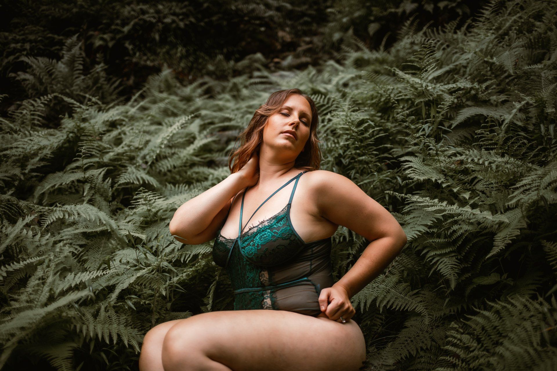 A woman in lingerie is sitting in the woods surrounded by ferns.