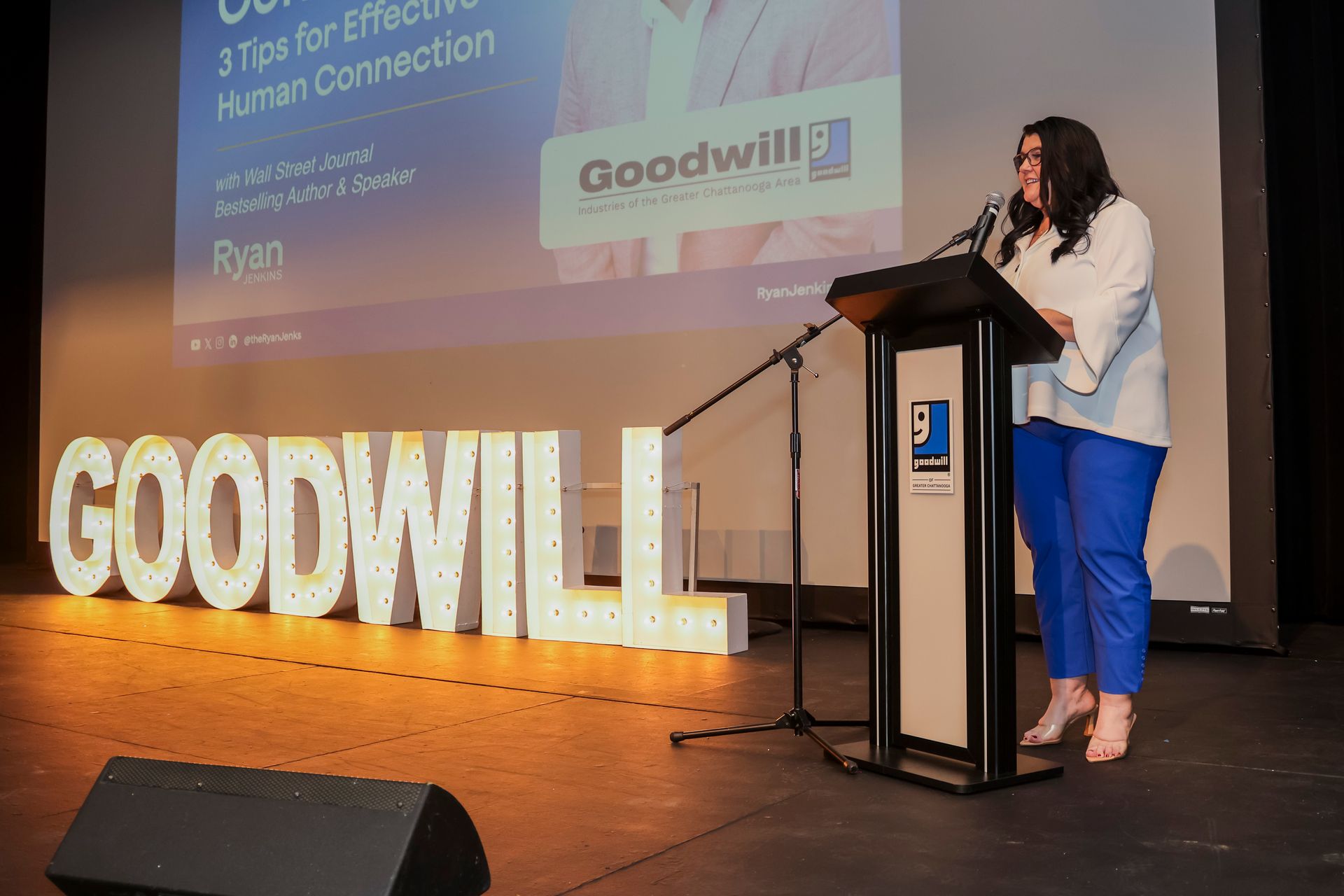 A person speaks at a podium in front of a large, illuminated 