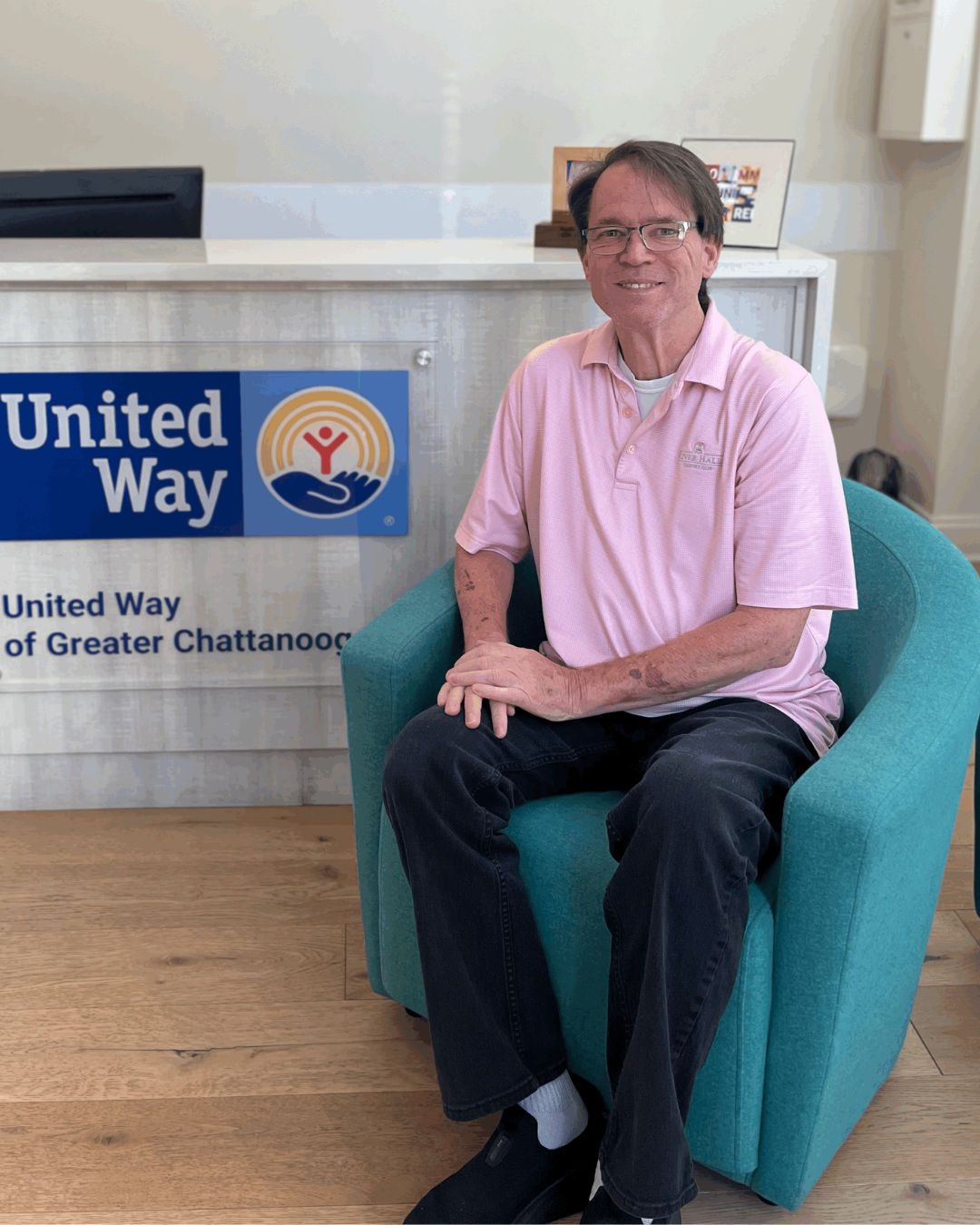 Man in pink shirt sits in blue chair in front of United Way sign.
