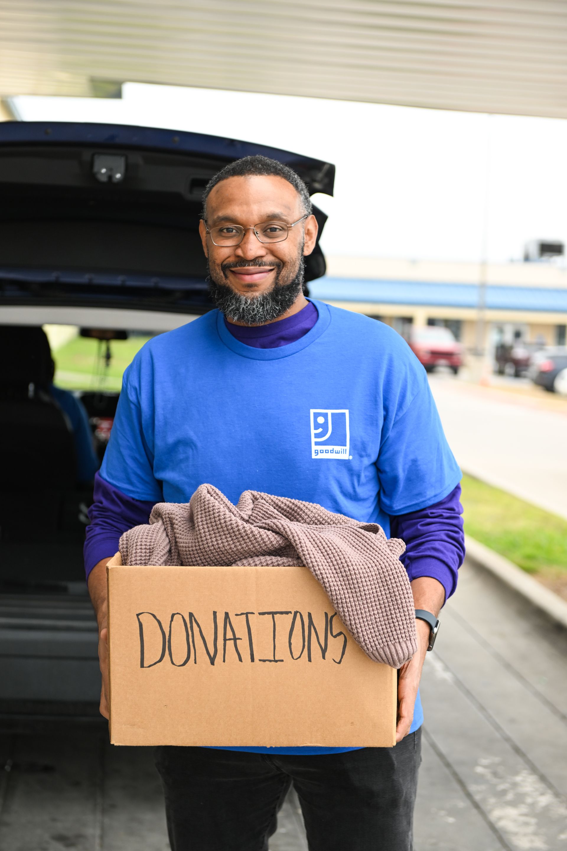 Un hombre sonríe y sostiene una caja de donaciones cerca del maletero abierto de un coche, con ropa encima.