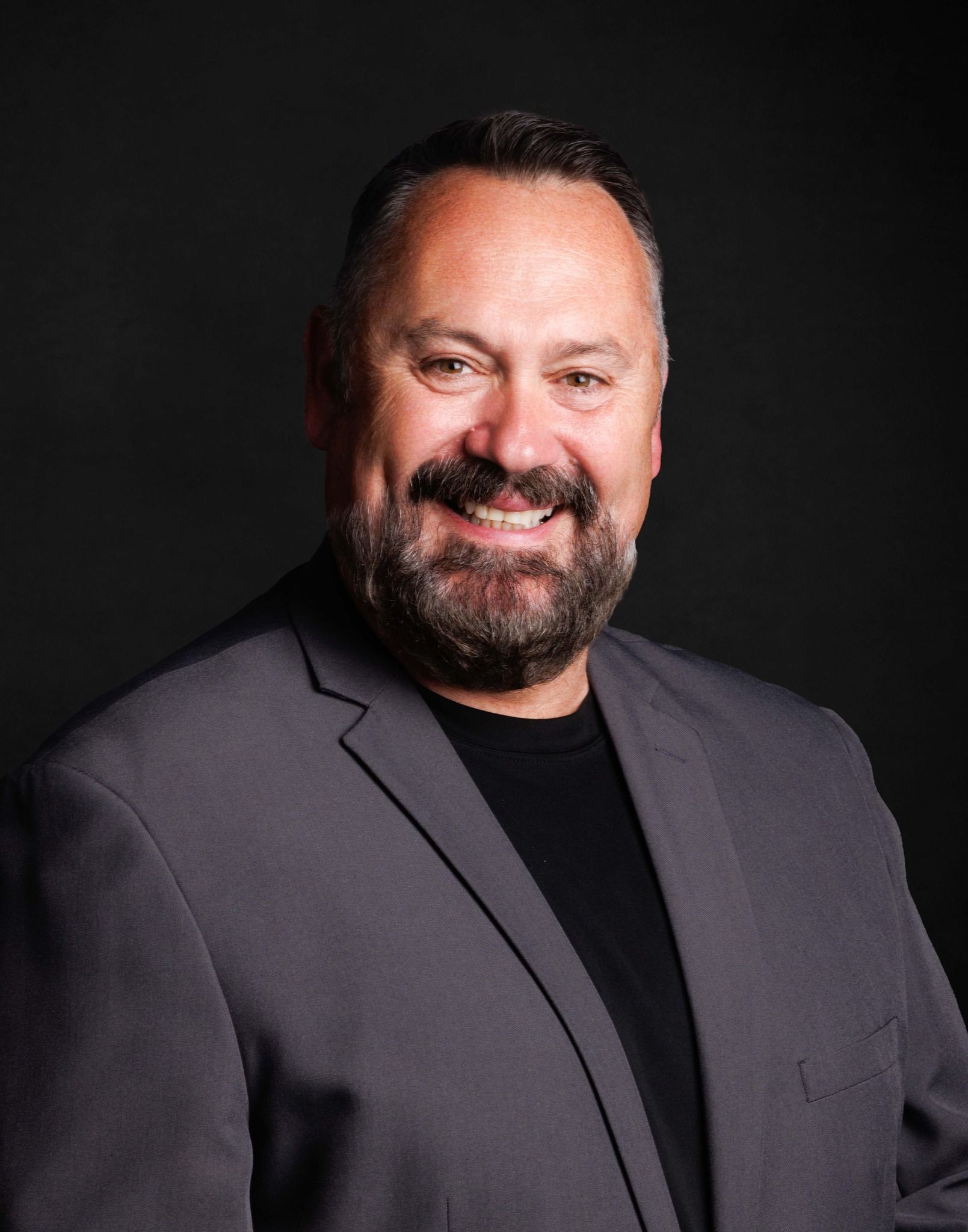Man in gray blazer smiles, black shirt, dark beard, against dark gray background.