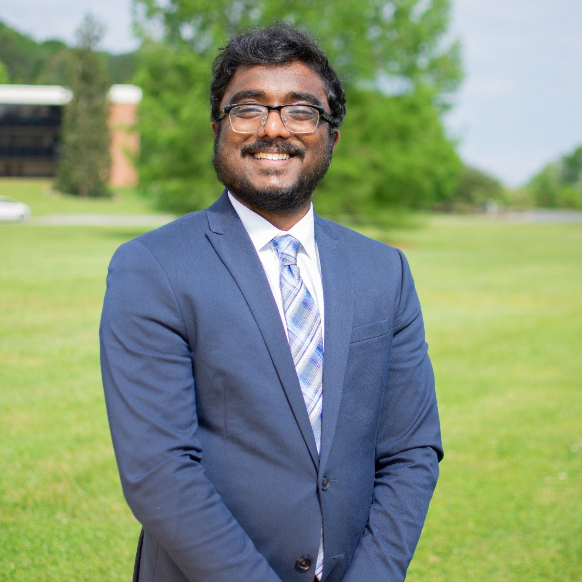 Man in blue suit and tie, smiling, standing on a grassy field with a building in the background.