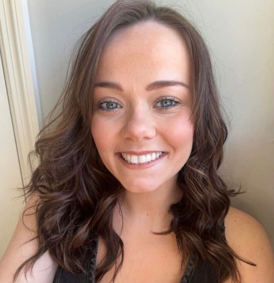 Woman with wavy brown hair smiles at the camera, wearing a dark top, against a neutral background.