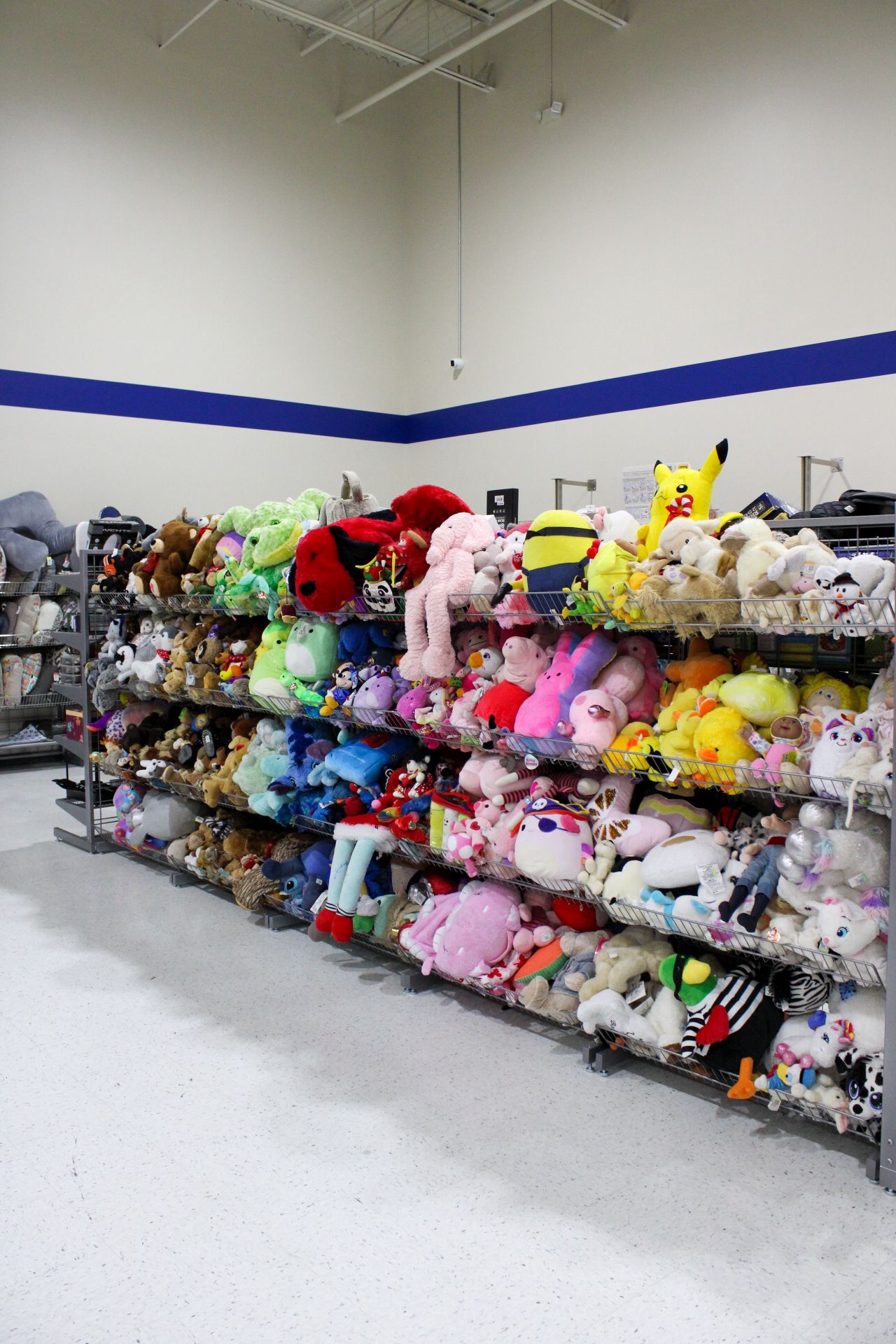 Shelves packed with various stuffed animals in a store. Bright colors dominate. White floor, blue wall stripe.
