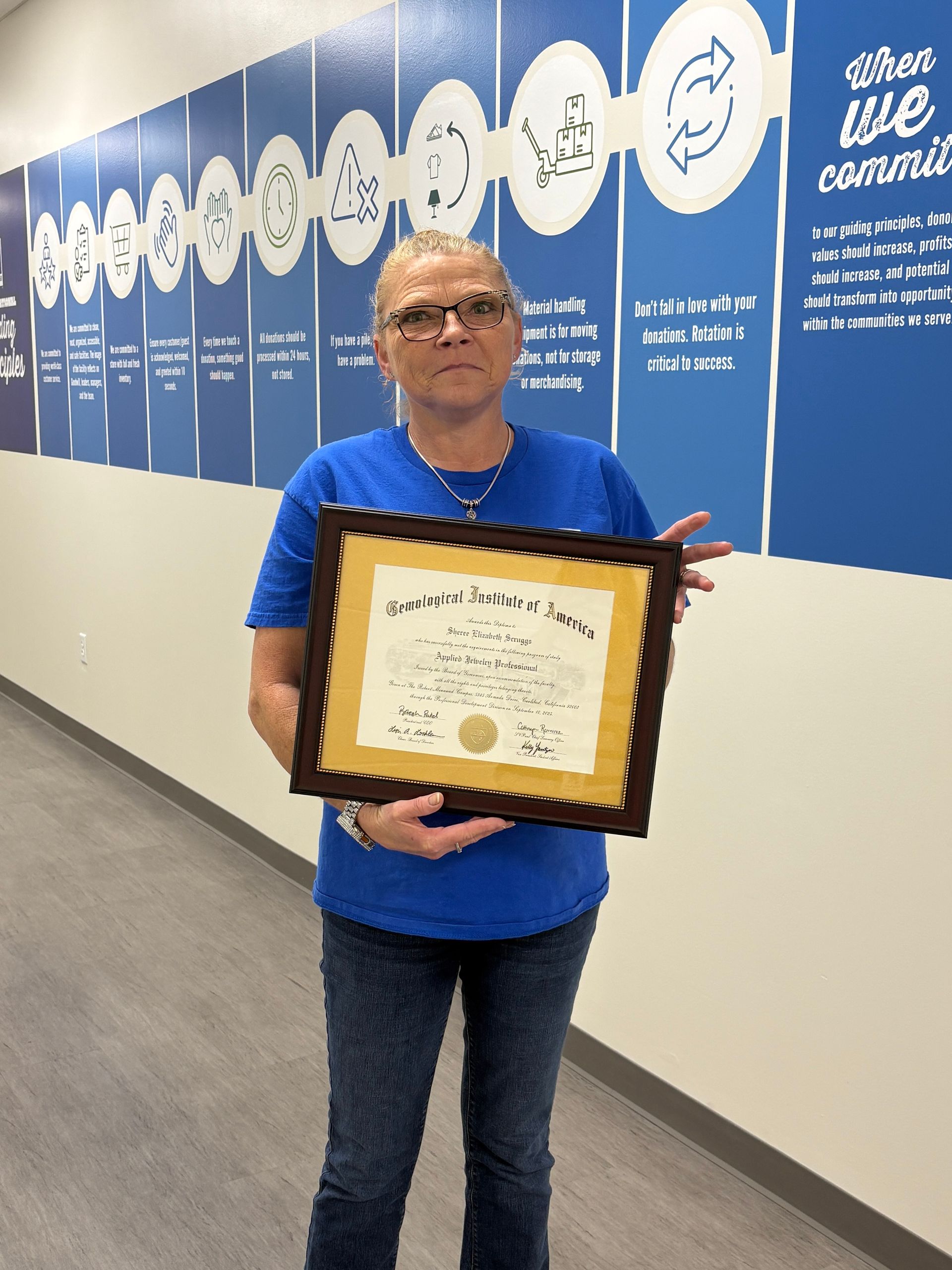 Woman in blue shirt and jeans holds a framed certificate in a hallway.