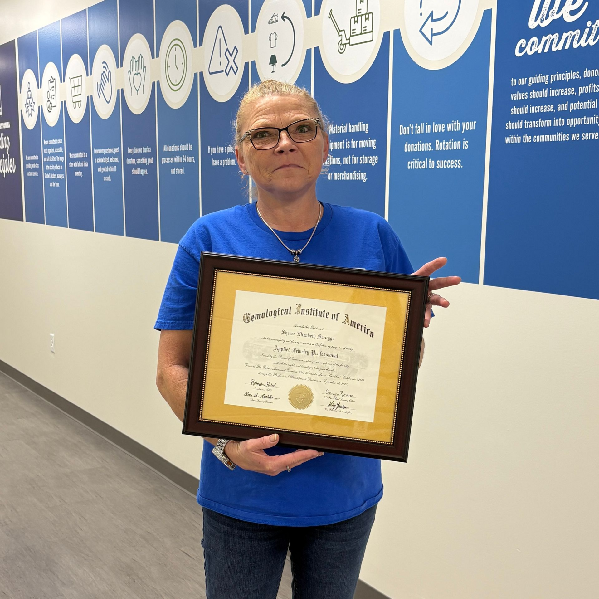Woman in blue shirt holding a framed certificate in a hallway with blue and white wall decorations.