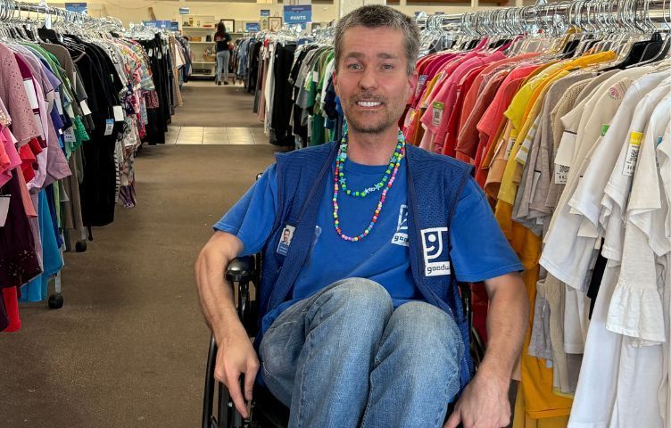Man in wheelchair at clothing store, smiling, wearing vest and beaded necklace, surrounded by racks of clothes.