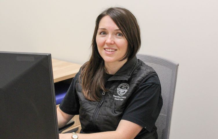 Woman in black vest and shirt smiles while sitting at a desk with a computer.