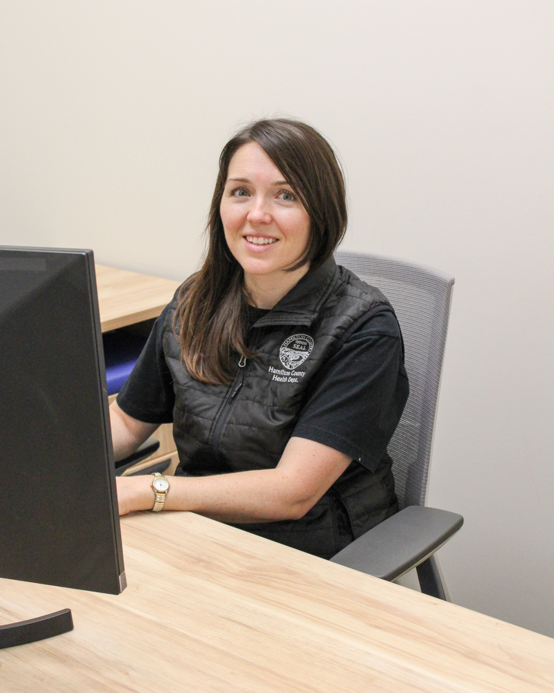 Woman in black vest and shirt smiles while sitting at a desk with a computer.