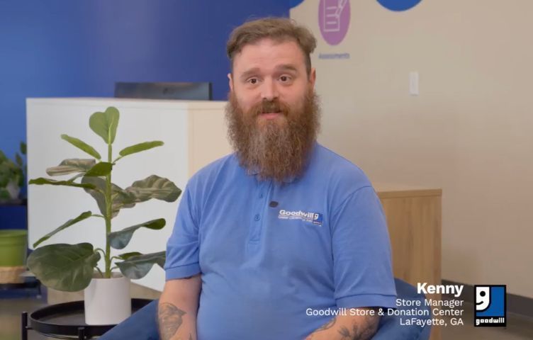 A bearded person in a blue polo shirt sits indoors next to a potted plant in front of a blue and beige wall.