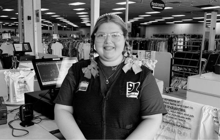 Woman with glasses and braids at a store checkout counter smiling in front of clothing racks.