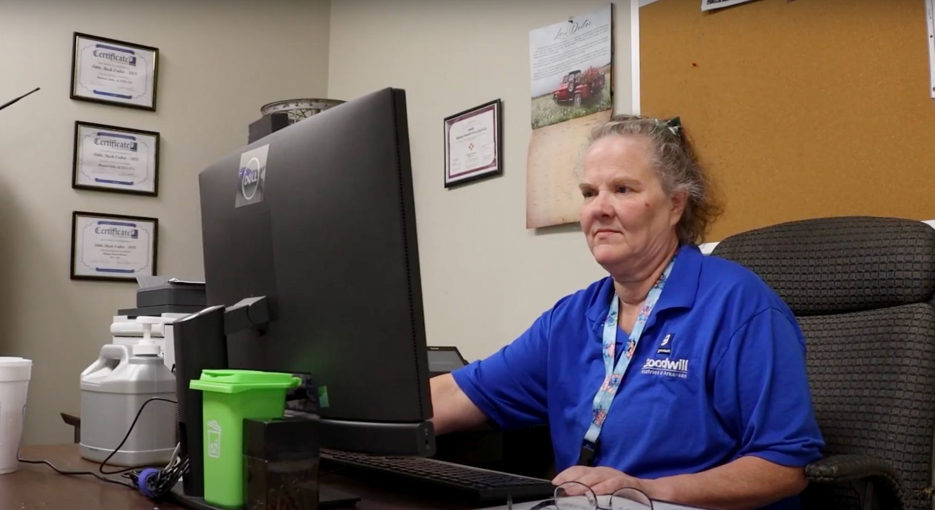 Woman in blue shirt seated at a desk with two computer monitors, typing. Office setting with certifications on the wall.