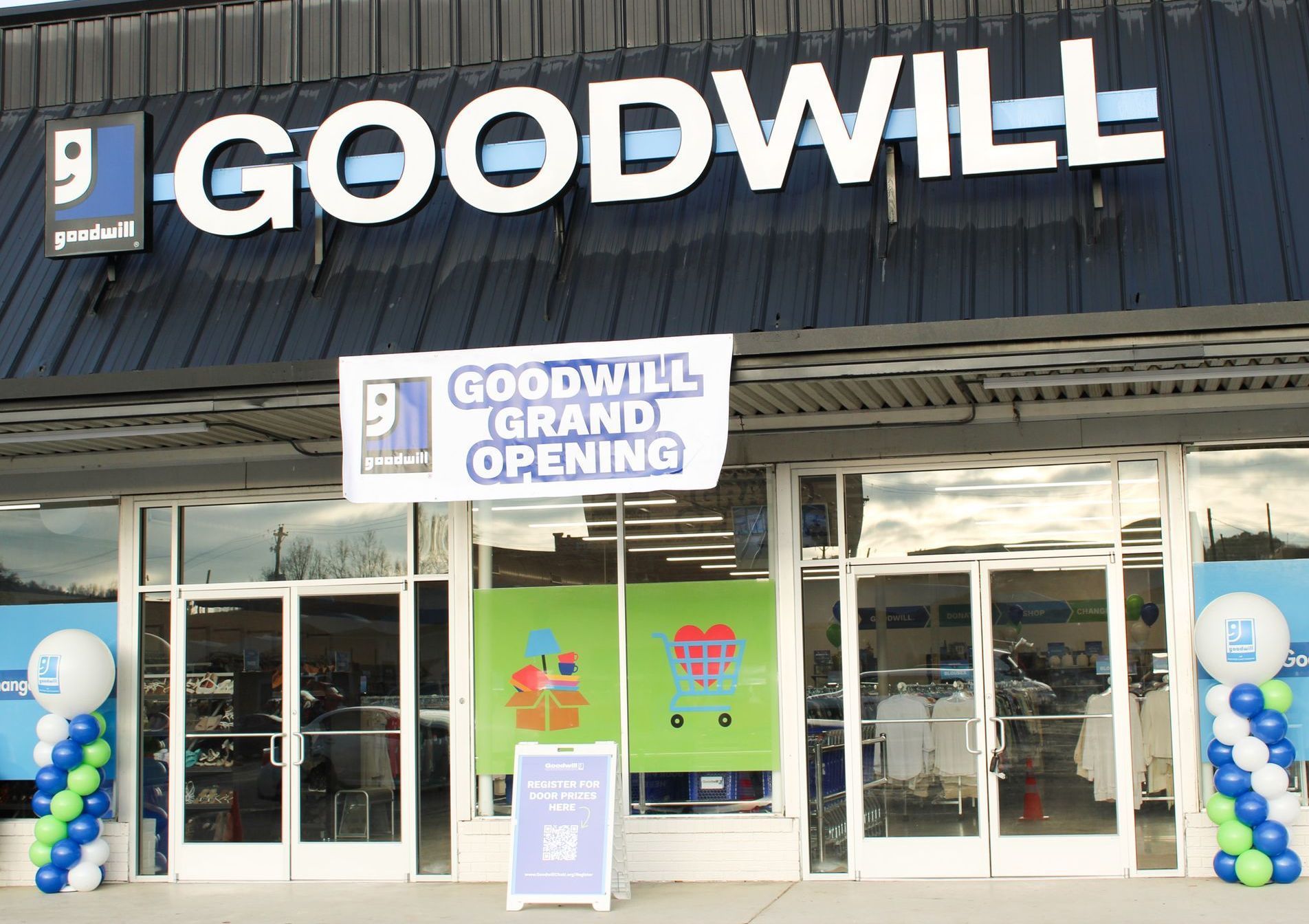 Fachada de una tienda Goodwill con letrero de gran inauguración, globos azules y blancos y carteles verdes en las ventanas.