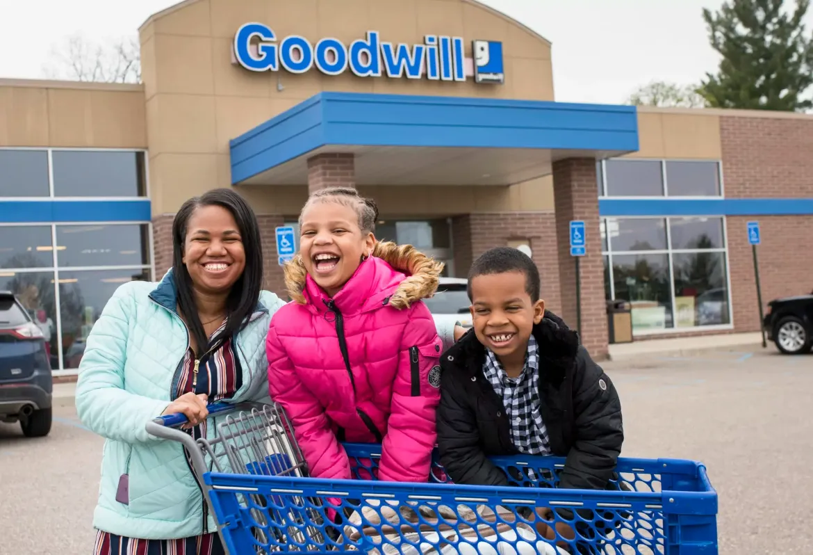 A woman and two children are pushing a shopping cart in front of a goodwill store.