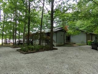 Grey-sided building surrounded by trees and a gravel driveway; covered porch on the left, car on the right.