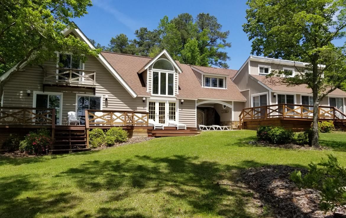 Beige and brown house with multiple levels, decks, and green lawn, surrounded by trees under a blue sky.