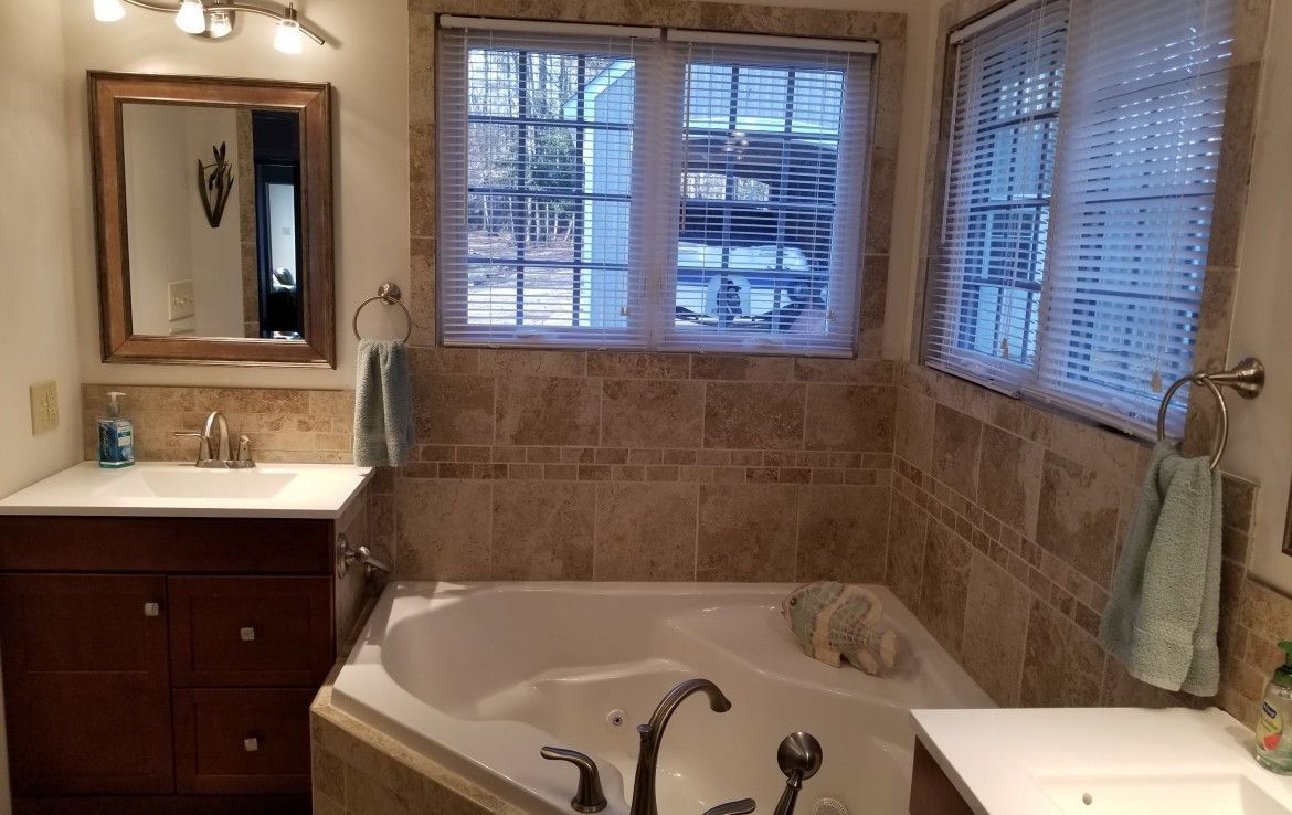 Bathroom with corner tub, two sinks, wood vanity, tiled walls, and windows with blinds.