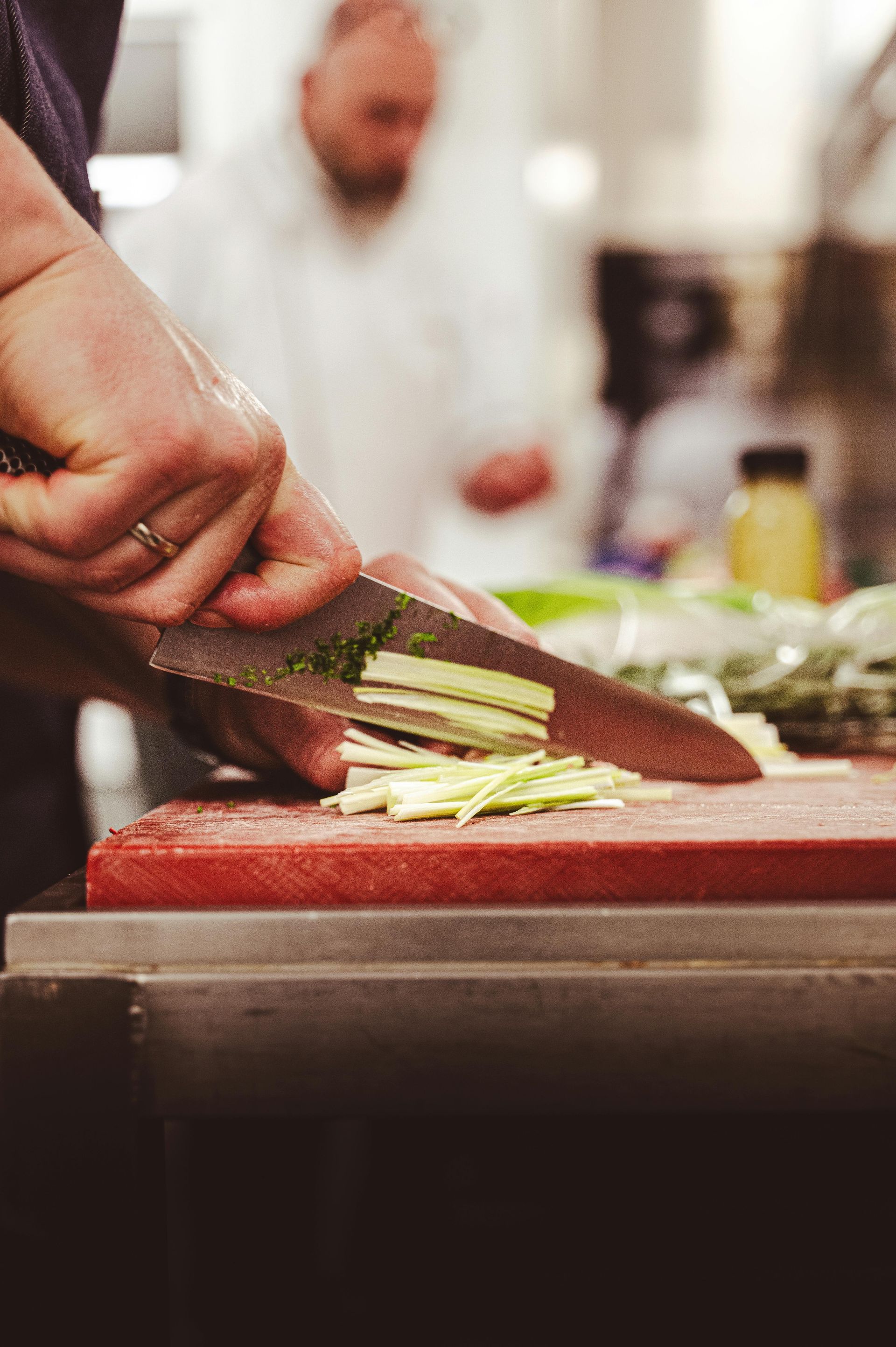 Hand slicing green vegetables on a red cutting board in a kitchen, with a blurred cook in the background