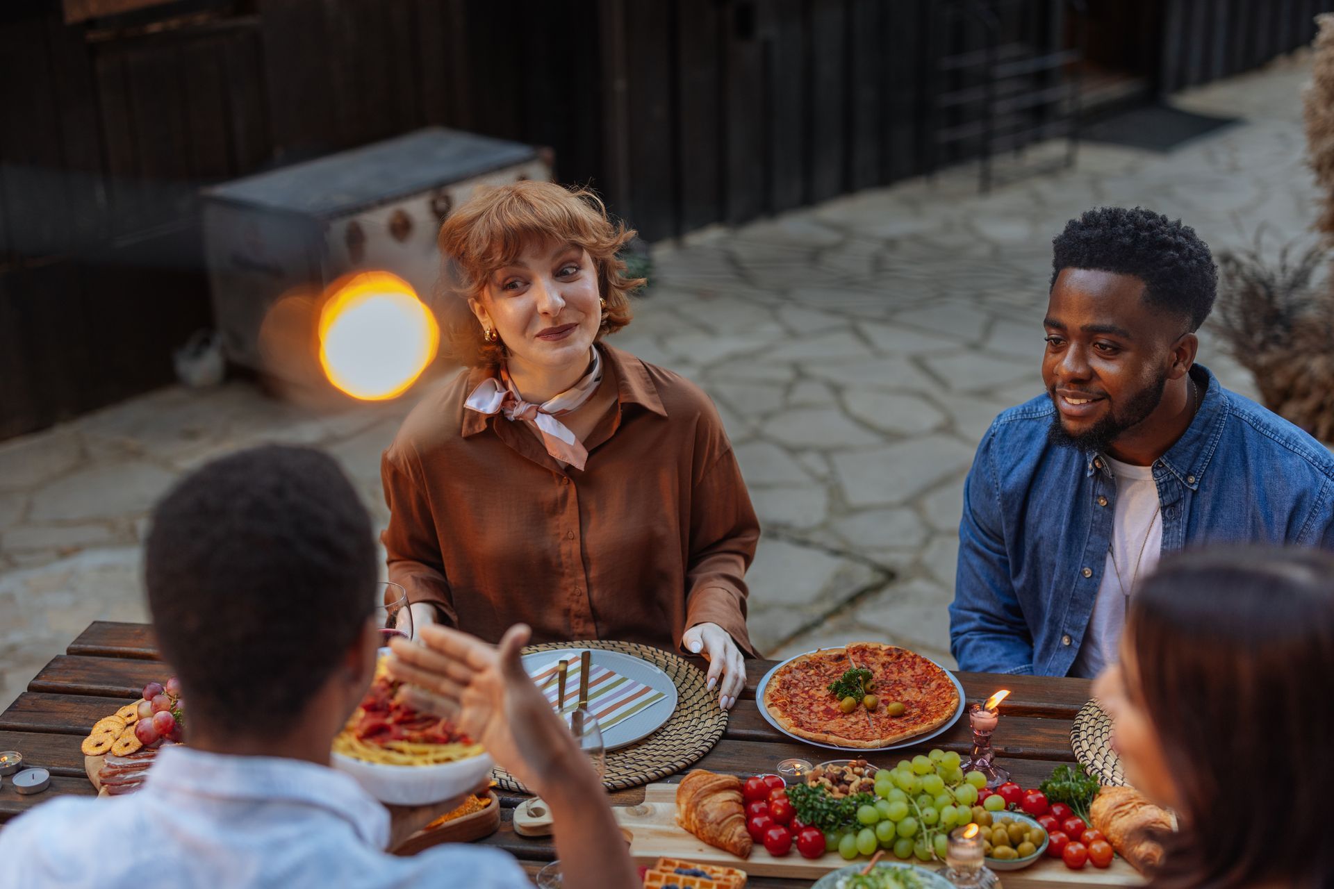 Friends chatting around a table with food outdoors near a glowing heater