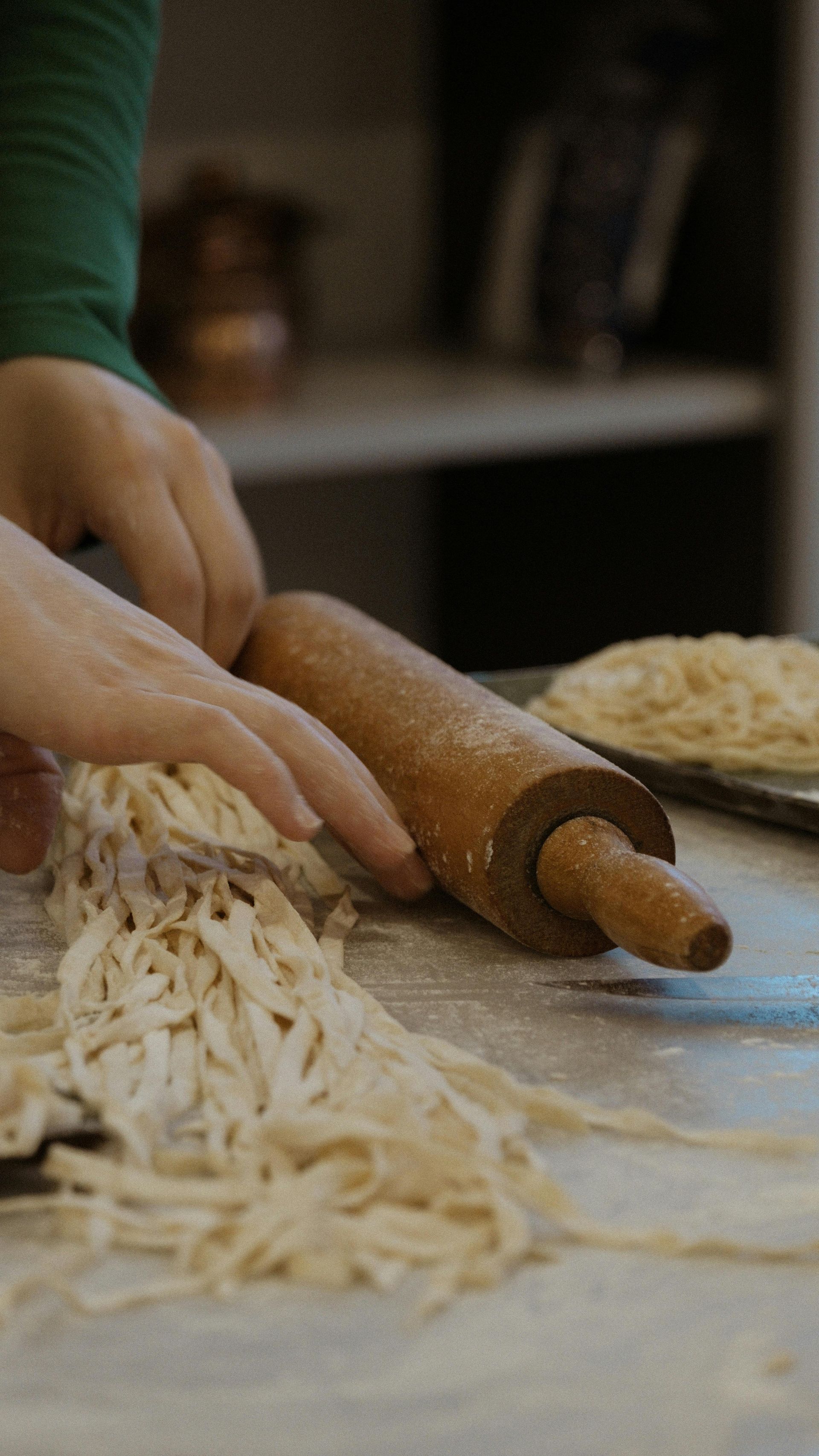 Hands rolling dough with a wooden pin beside a pile of fresh noodles in a kitchen