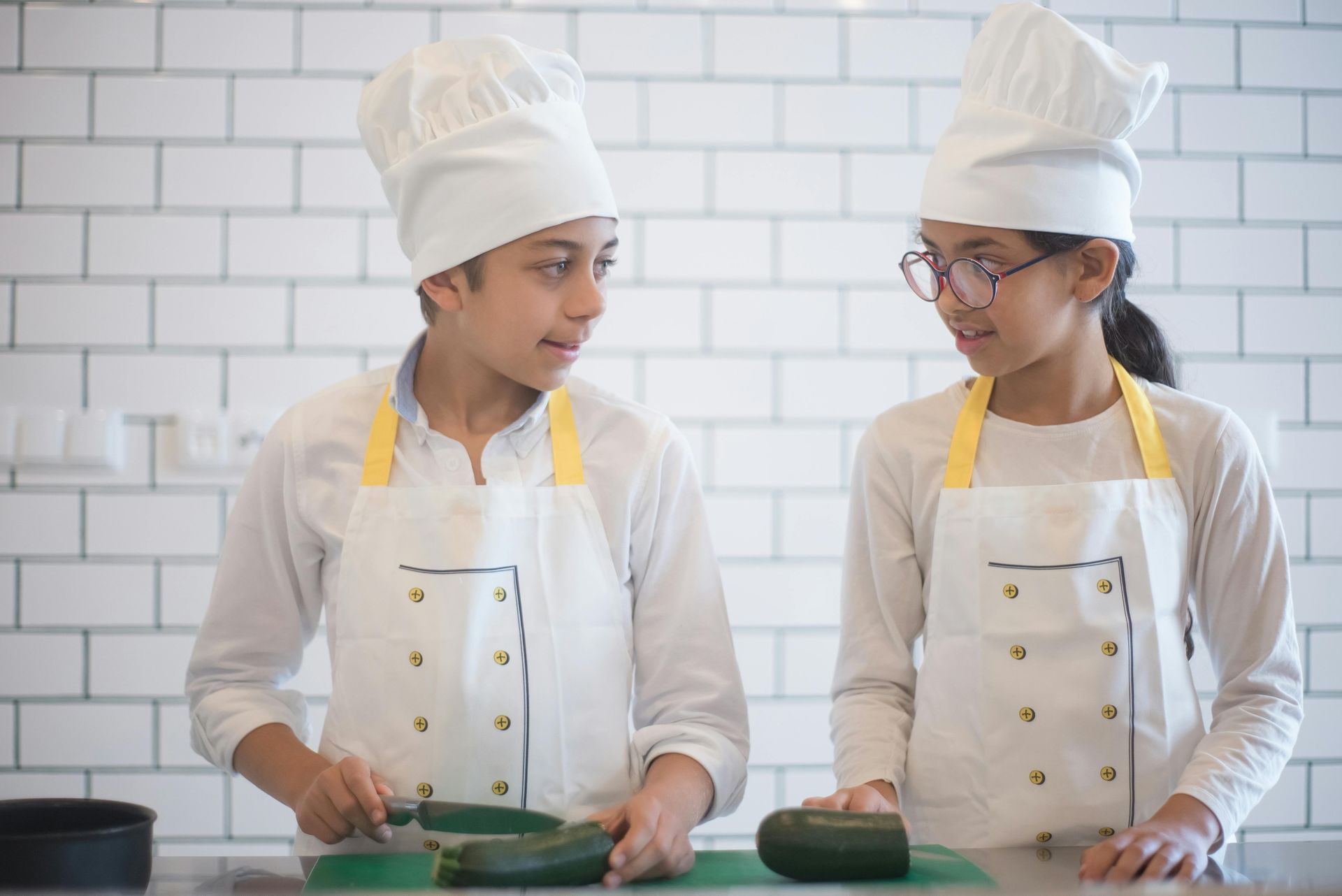 Two chefs in white uniforms and hats preparing green dough in a tiled kitchen