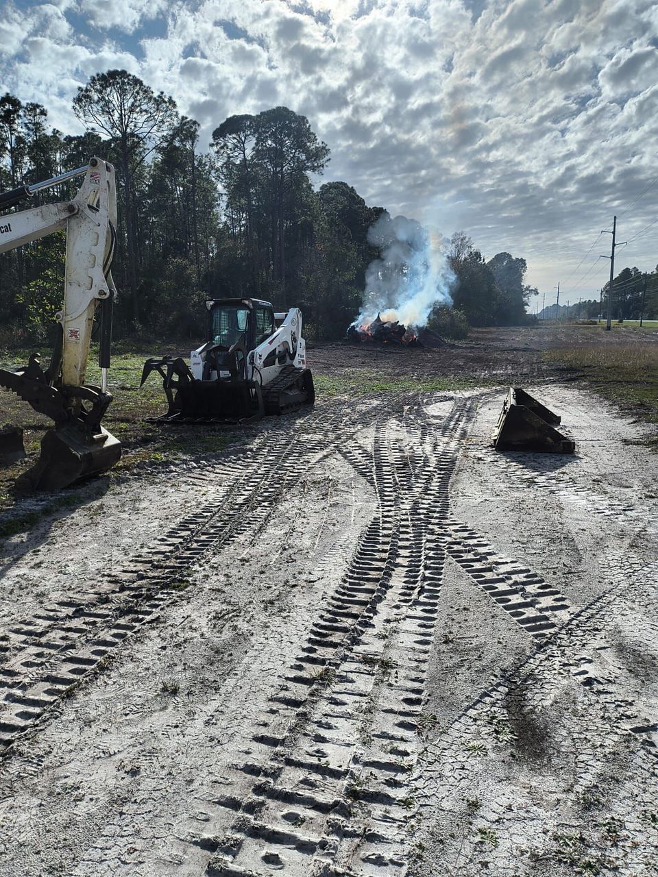 Construction site with muddy tire tracks, a small excavator, and smoke rising in the background