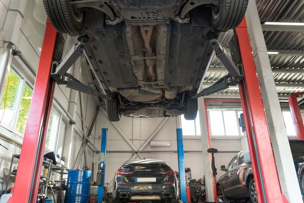 Car on a lift in a repair shop. Red lift posts, blue equipment, other cars visible.